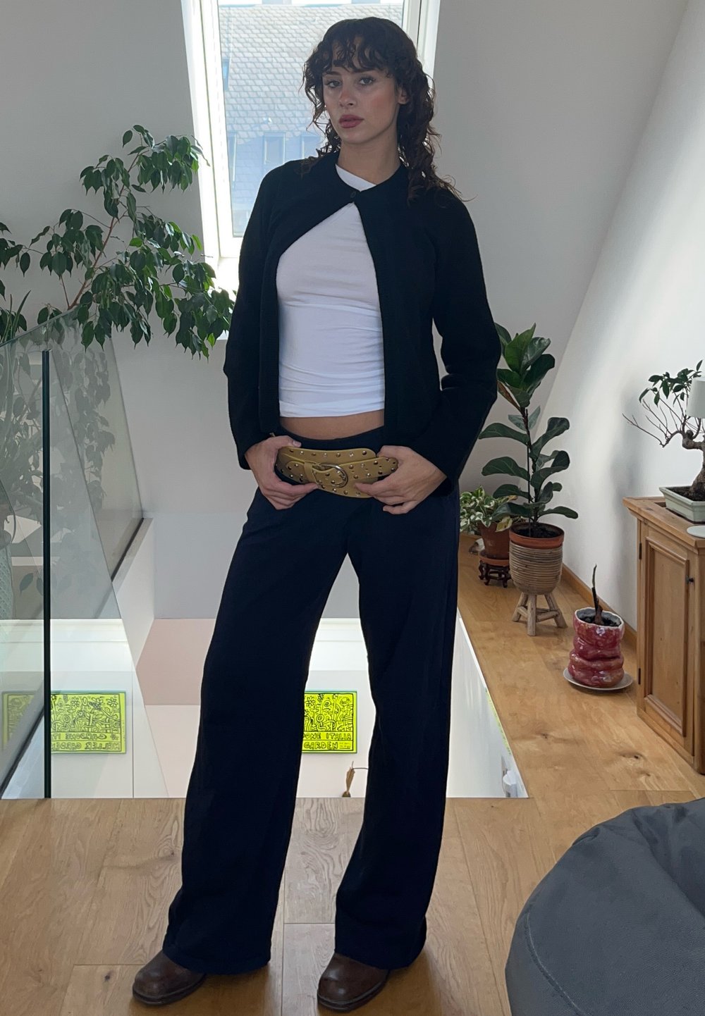 Μαύρο κοντό καρντιγκάν, άσπρο εφαρμοστό πουκάμισο, φαρδιά ναυτικά παντελόνια, χρυσή ζώνη με καρφιά, καφέ παπούτσια. Εσωτερικός χώρος με φυτά και ξύλινο πάτωμα.