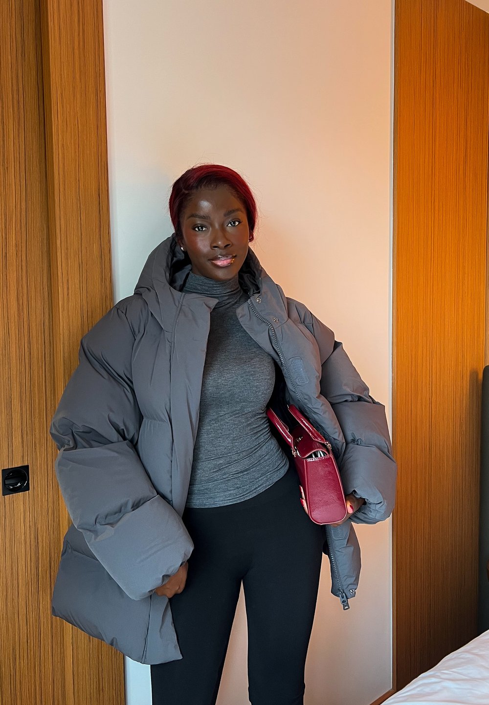 Femme aux cheveux bordeaux portant une grande doudoune grise, un col roulé gris, un pantalon noir, tenant un sac à main rouge, debout à l'intérieur près de panneaux en bois.