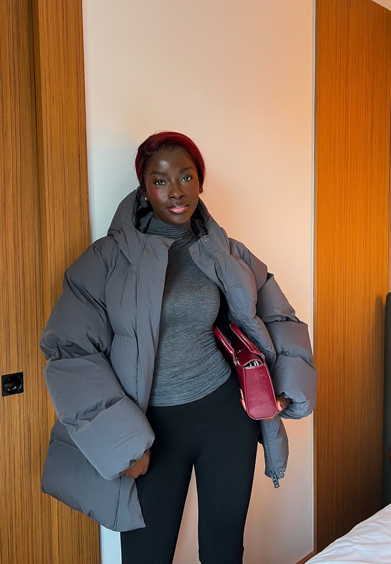 Mujer con cabello burdeos que lleva una gran chaqueta acolchada gris, cuello alto gris, pantalones negros y sostiene un bolso rojo, de pie en el interior junto a paneles de madera.