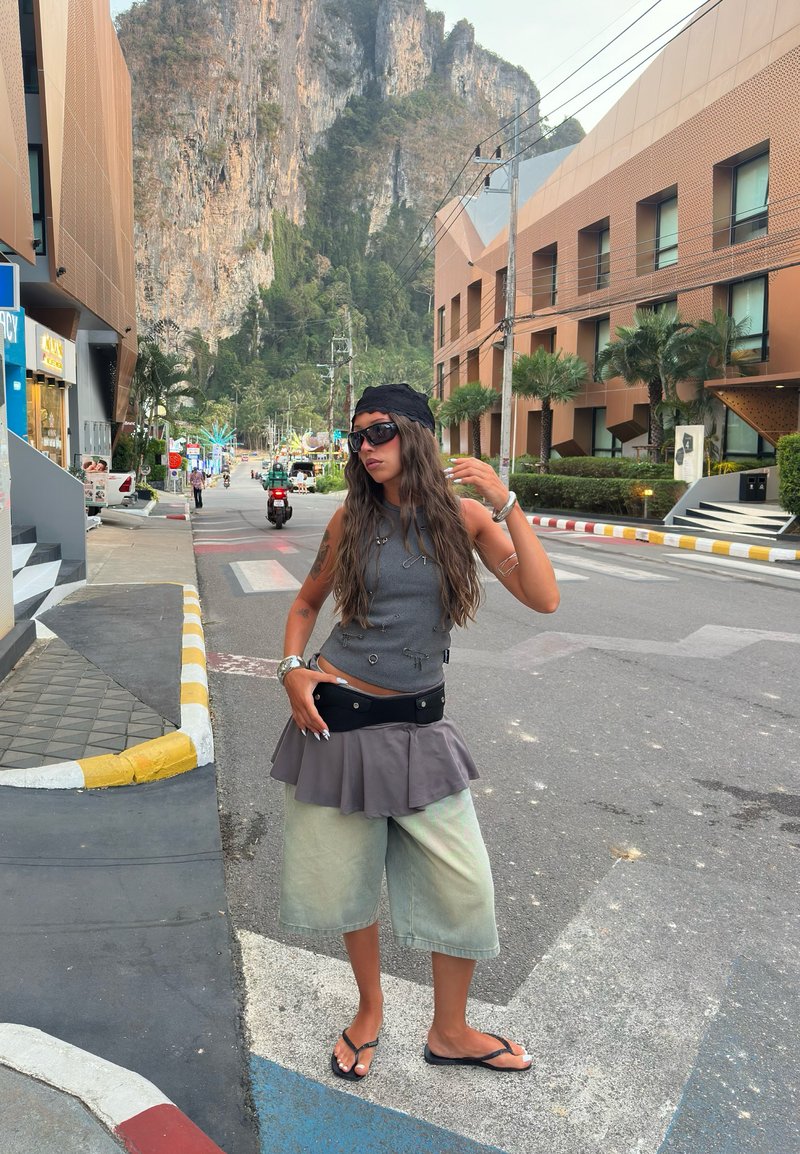 Γυναίκα με γυαλιά ηλίου και καθημερινά ρούχα στέκεται στο δρόμο με ψηλό βραχώδη γκρεμό και σύγχρονα κτίρια στο παρασκήνιο.