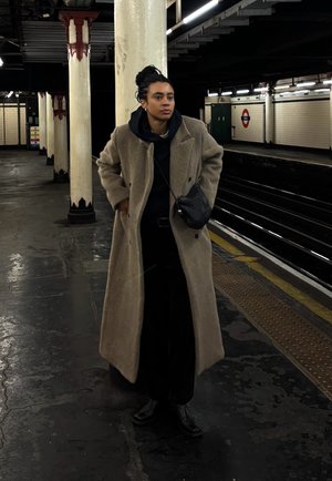 Persona con abrigo largo beige y sudadera con capucha negra de pie en el andén del metro cerca de las vías, con columnas blancas y paredes de azulejos al fondo.