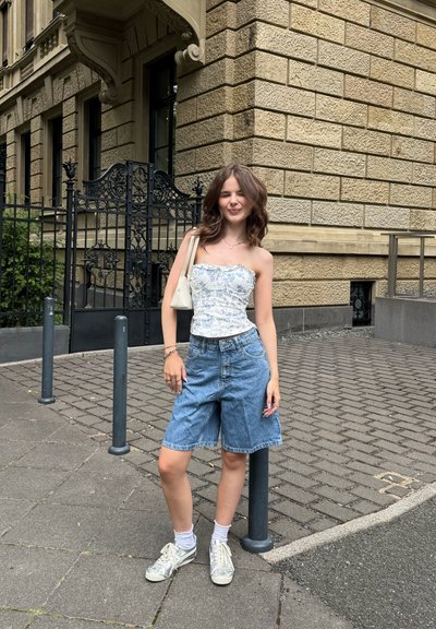 Top corset de patrón floral en azul claro, combinado con pantalones cortos de mezclilla holgados y zapatillas grises. Ambientado frente a un edificio de piedra y una barandilla de metal.