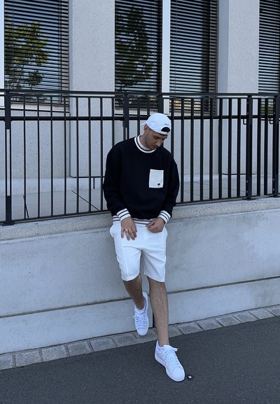 Sudadera negra con puños y bolsillo a rayas blancas, pantalones cortos blancos y zapatillas blancas. De pie contra una pared de concreto.