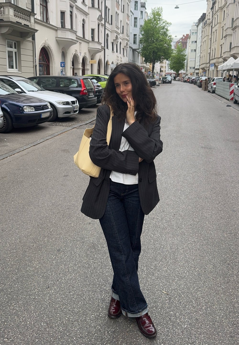 Blazer noir à rayures, porté sur une chemise blanche, un jean large foncé et des chaussures bordeaux. Porte un sac fourre-tout jaune clair. Cadre urbain.