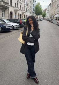 Blazer noir à rayures, porté sur une chemise blanche, un jean large foncé et des chaussures bordeaux. Porte un sac fourre-tout jaune clair. Cadre urbain.