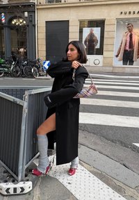 Kvinne i svart frakk og grå leggvarmere justerer en rutete veske på fortauet i byen ved et fotgjengerfelt, med motebutikkplakater bak.