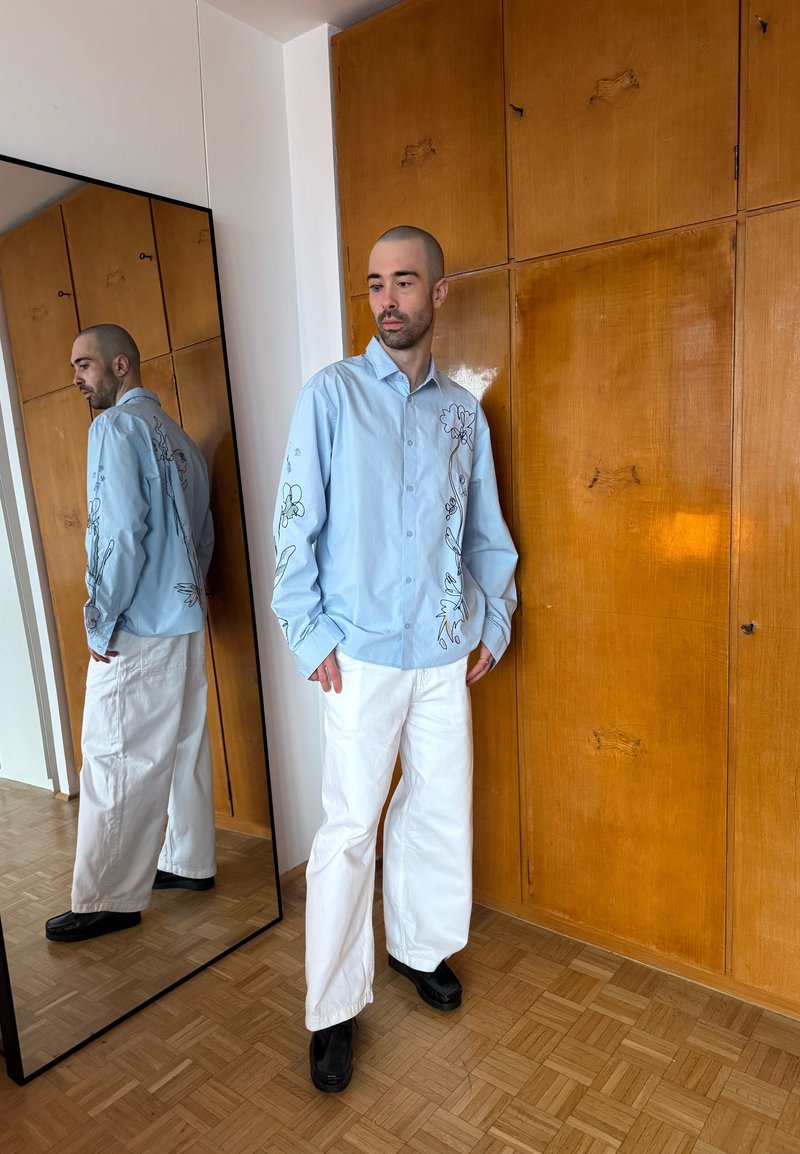 Homme portant une chemise bleu clair avec broderies florales, pantalon blanc et chaussures noires, debout à côté d'une armoire en bois et d'un grand miroir.
