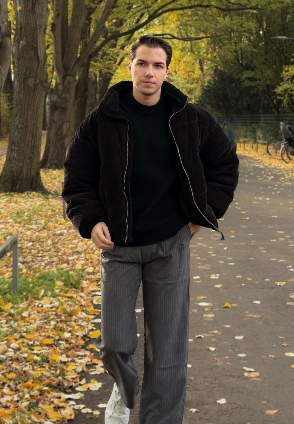 Fekete steppelt dzseki színes cipzárral, bordázott zöld pulóver és szürke csíkos nadrág. Fehér sportcipő. Őszi levelek a háttérben.