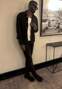 Czarna kurtka dżinsowa na białym swetrze z dekoltem w serek, zestawiona z czarnymi dżinsami i butami wsuwanymi, stojąca obok ściany z oprawionym zdjęciem.