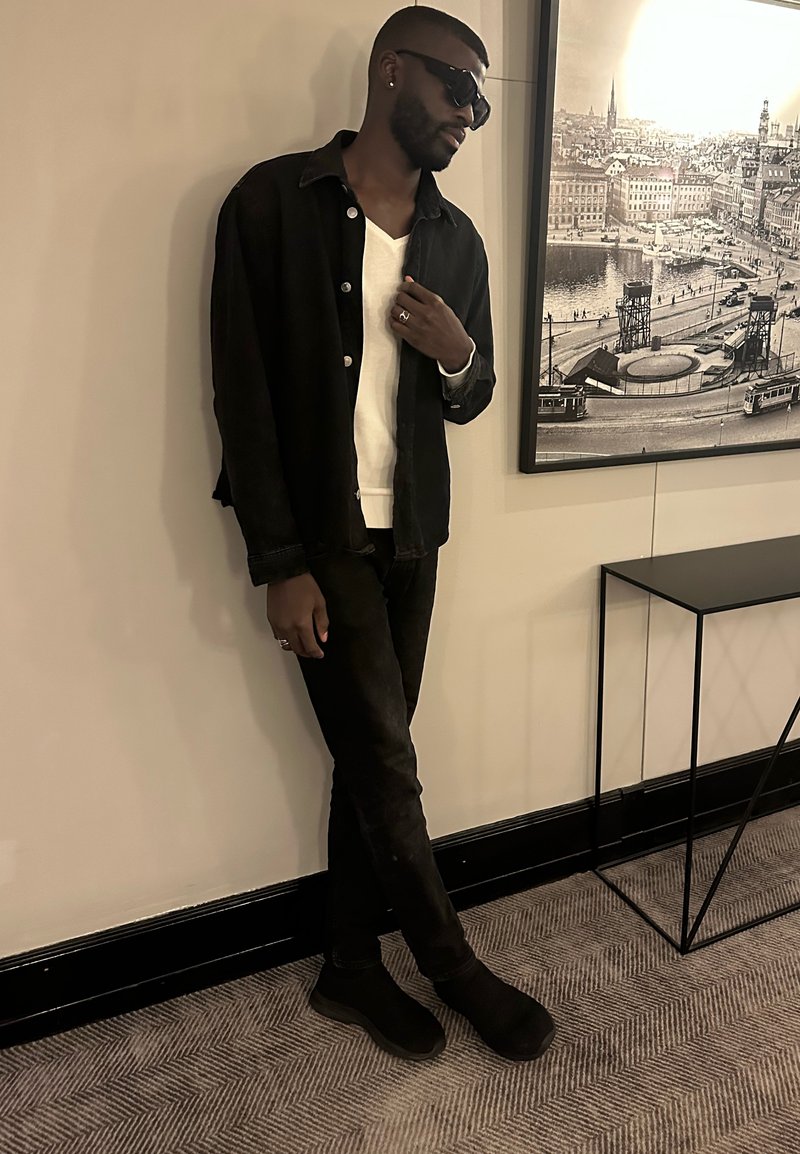 Giacca di denim nera su un maglione bianco a scollo a V, abbinata a jeans neri e scarpe slip-on, in piedi accanto a un muro con una fotografia incorniciata.