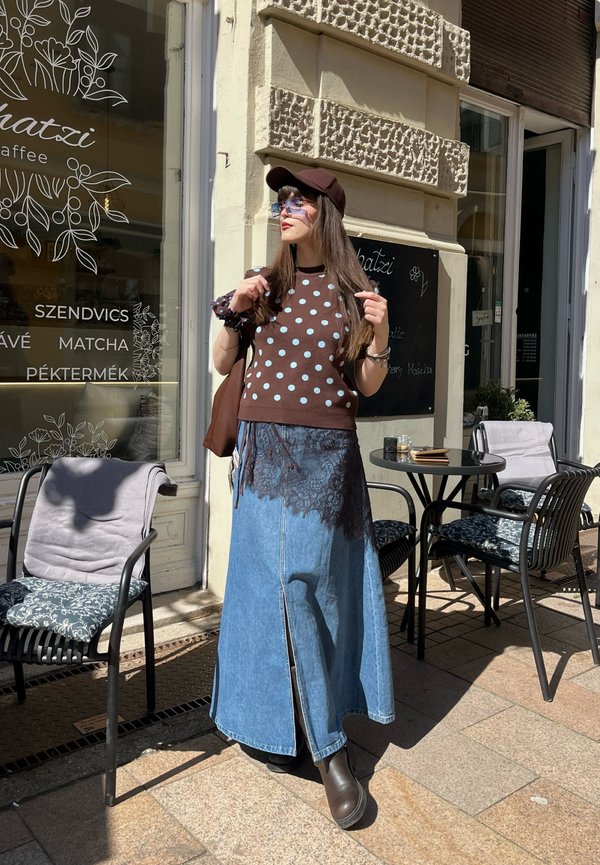 Kvinde iført brun polkaprikket sweater, blå denimnederdel med sort blonder, brun kasket og solbriller, står ved caféborde udenfor.