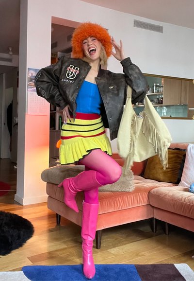 Mujer con sombrero de piel naranja brillante, blusa azul, chaqueta negra, falda amarilla, medias rosas y botas, sosteniendo un bolso beige con flecos en una sala de estar colorida.