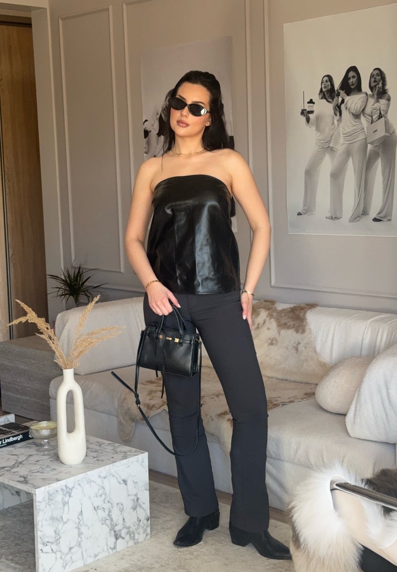 Femme portant un haut noir sans bretelles et un pantalon, lunettes de soleil, tenant un sac à main noir, debout dans un salon moderne avec une œuvre d'art monochrome sur le mur.