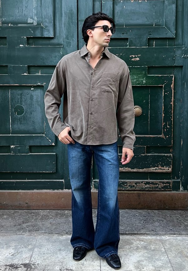 Hombre con gafas de sol, camisa gris suelta abotonada y vaqueros anchos azules, de pie frente a una gran puerta de madera verde con pintura descascarada.