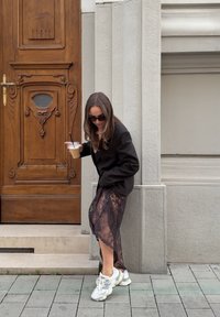 Čierny blazer a priesvitná čierna čipkovaná sukňa, skombinované s bielymi teniskami. Drží nápoj a opiera sa o drevené dvere s podrobnými rezbami.