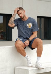 Maglia sportiva grigia con logo "Los Angeles Lakers", indossata con pantaloni corti coordinati. Presenta un design con bottoni e scarpe da ginnastica bianche indossate con calze.