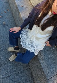 Mujer sentada en unos escalones de piedra, vestida con una chaqueta oscura, blusa blanca de encaje, vaqueros azules y zapatos beige con calcetines negros.