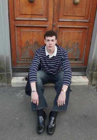 Camisa polo de rayas en azul marino y blanco de manga larga, jeans oscuros, mocasines negros brillantes, sentado en una acera frente a puertas de madera.