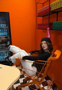 Chariot de magasinage métallique orange avec un design en fils, doté de roues noires. Les murs en arrière-plan sont d'un orange vif ; les étagères exposent des canettes de boissons colorées.