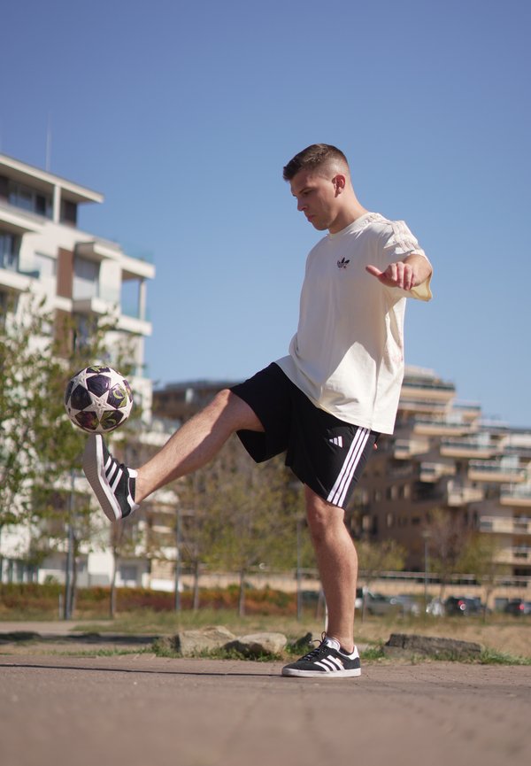 Joven con camisa blanca y pantalones cortos negros haciendo malabares con un balón de fútbol al aire libre, con edificios urbanos y cielo despejado de fondo.