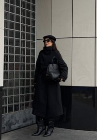 Lang svart kåpe med høy krage og belte, kombinert med svarte hansker, strukturert håndveske og spisse støvler. Mørk caps og solbriller.