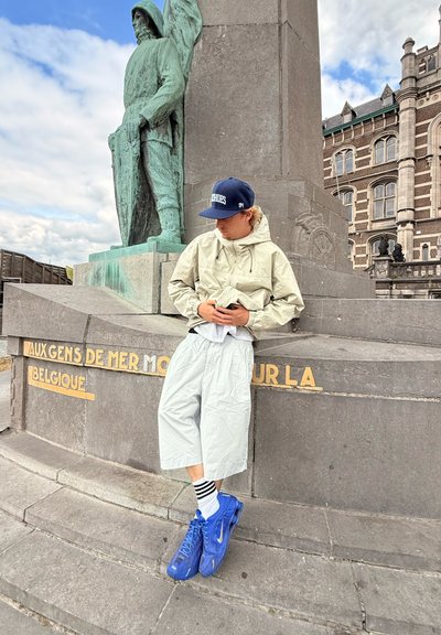 Rompevientos gris claro con capucha, pantalones cortos holgados a juego y zapatillas azules con rayas blancas. Estatua de una figura en el fondo.