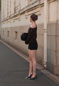 Czarny sweter o luźnym splotcie, czarne szorty i czarne szpilki ze złotymi końcówkami. Brązowe włosy spięte w kok, trzyma czarną torebkę przy ścianie.