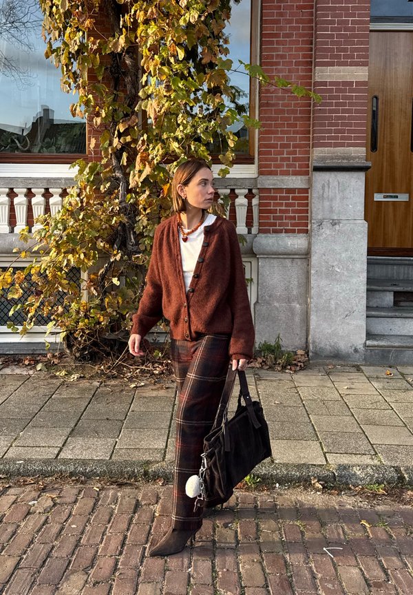 Brun, strukturert cardigan med knapper, rutete bukser i mørke toner, og en svart skulderveske. Fortausbakgrunn med høstløv.