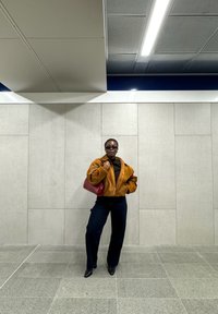 Veste en suédine marron avec fermeture éclair, chemise noire, pantalon sombre et escarpins noirs. Tient un sac à main rouge. Fond carrelé gris.