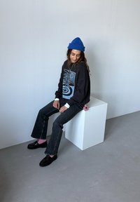 Sweat-shirt noir oversize avec un motif graphique bleu, associé à un jean évasé foncé et des chaussures noires, avec des chaussettes roses et un pendentif en forme de fleur.