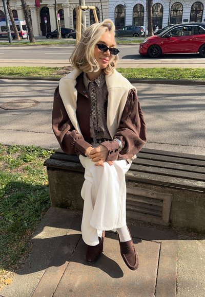 Mujer con cabello rubio que lleva gafas de sol, camisa marrón, pantalones blancos y una chaqueta crema colocada sobre los hombros, sentada en un banco del parque a la luz del sol.