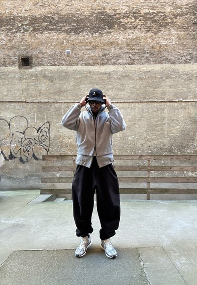 Sudadera gris con cremallera, pantalones de pierna ancha negros y zapatillas plateadas. Los accesorios incluyen un sombrero tipo cubo negro con diseño de calavera y gafas de sol.