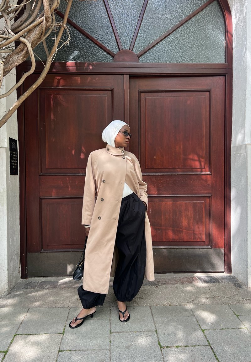 Femme portant un trench-coat beige, un foulard blanc, des lunettes de soleil noires et un pantalon noir ample, debout devant de grandes portes doubles en bois.