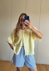 Κίτρινο, ριχτό cardigan με κοντά μανίκια, κλείσιμο με κουμπιά, φορεμένο πάνω από ένα ανοιχτό τοπ και ριγέ μπλε σορτς. Λείος υφασμάτινος χαρακτήρας, άνετη εφαρμογή.