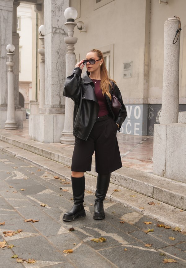 Una mujer con gafas de sol, una chaqueta de cuero negra, pantalones cortos y botas hasta la rodilla se ajusta las gafas en un paseo urbano empedrado.