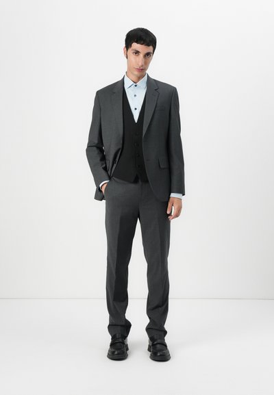 Traje gris de corte ajustado, con solapas de pico y cierre de un solo botón. Lleva una camiseta de vestir azul clara debajo del chaleco negro.