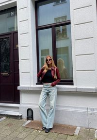 Červená kožená bunda, černý top, světle modré široké džíny, černé boty. Stojí opřený o texturovanou bílou zeď poblíž okna a dveří.