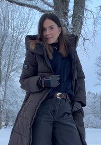 Femme en manteau d'hiver noir et gants, debout en plein air dans un paysage enneigé avec des arbres dépouillés.