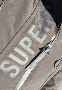 Zwarte stoffen tas met grote, reliëf "SUPERDRY" tekst. Inclusief een zwarte rits met een logo-trekhaak en een gladde textuur.