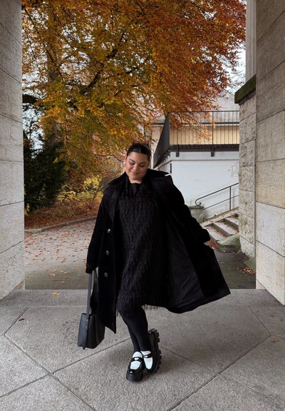 Abrigo de lana negro sobre un vestido negro con flecos, combinado con medias negras y zapatos negros gruesos con detalles blancos. Bolso negro en la mano.
