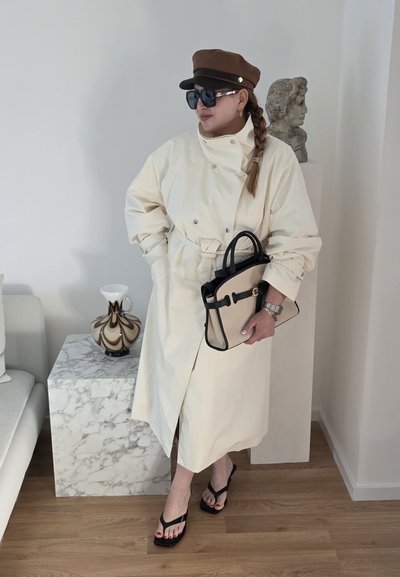 Mujer con gabardina beige, gorra marrón, gafas de sol negras, sosteniendo bolso beige y negro, de pie en el interior cerca de una mesa de mármol con jarrón y busto escultórico.