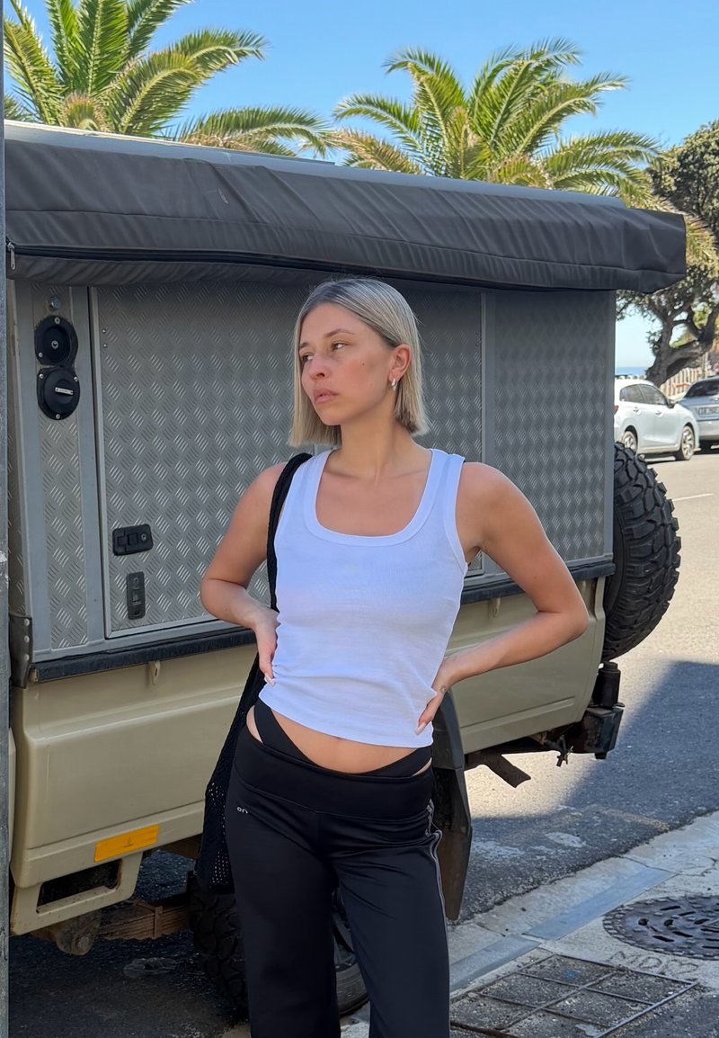 Young woman in white tank top and black pants stands with hands on hips beside beige vehicle with palm trees in background.