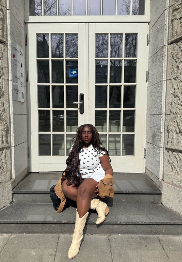 Žena s dlhými vlasmi, oblečená v bielom tričku s bodkami, bielej sukni a béžových čižmách, sedí na kamenných schodoch pred sklenenými dvojkrídlovými dverami.