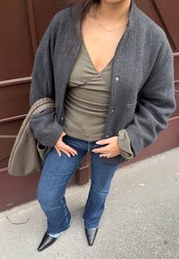 Grå ull bomberjakke, olivengrønn tettsittende topp, mørkeblå vide jeans og svarte spisse støvler, holder en brun vevd veske.