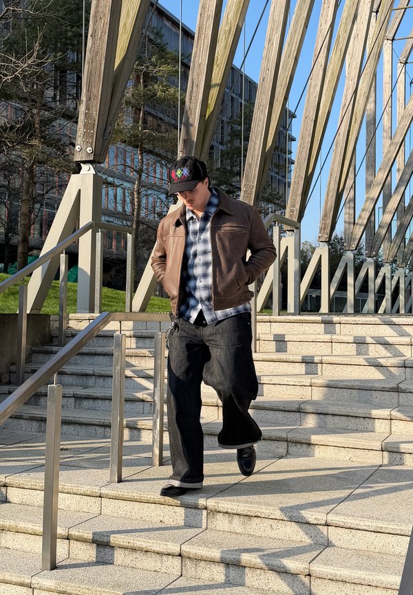 Fiatal férfi barna dzsekiben és fekete sapkában, aki kültéri kőlépcsőn sétál, mely mellett fém korlát van, és fölötte fa gerendák húzódnak.