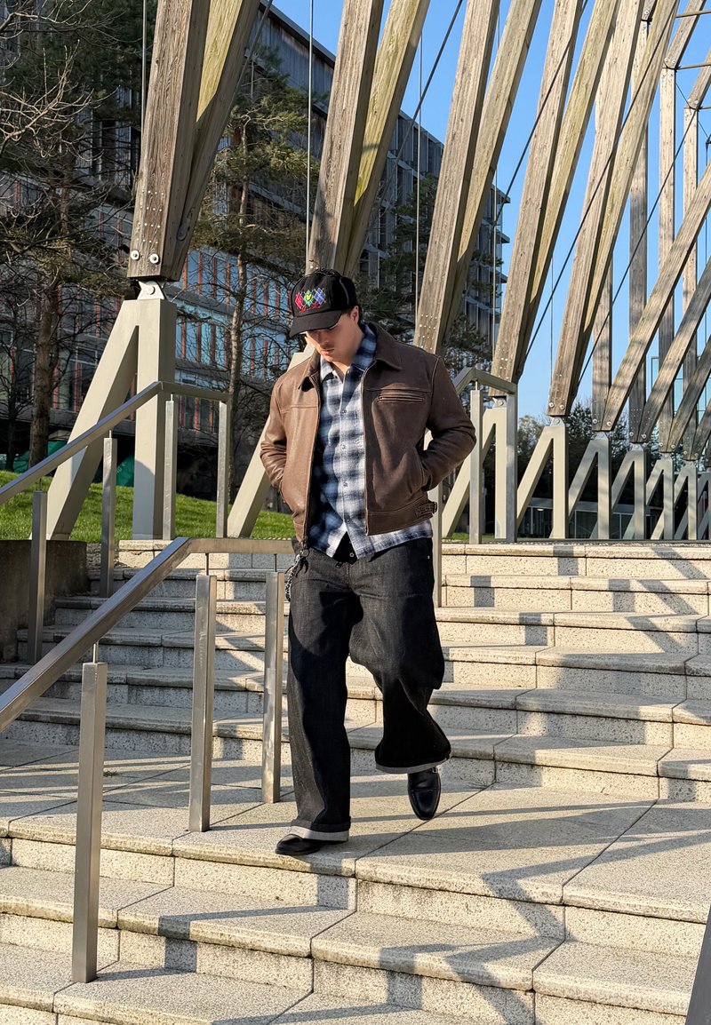 Joven con chaqueta marrón y gorra negra caminando por unas escaleras de piedra al aire libre, con pasamanos de metal y vigas de madera encima.