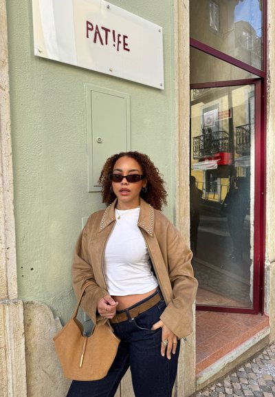 Chaqueta de ante marrón con cuello studded, camisa blanca y jeans oscuros. Sosteniendo un bolso de ante color beige. El fondo presenta un cartel de tienda y el reflejo de la ventana.