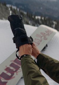 Sorte vanntette hansker med teksturert håndflate og borrelås, brukt mens man justerer et snowboard på snødekt terreng.
