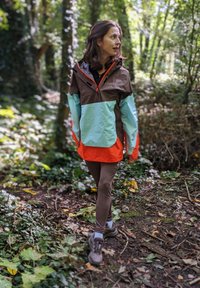 Lysfarvet jakke med brune, teal og orange paneler; kombineret med brune leggings og sko, placeret i et skovområde med løv.
