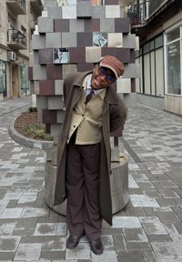 Άνδρας με καφέ παλτό, μπεζ πουκάμισο και σκούρα παντελόνια στέκεται δίπλα σε μια γεωμετρική γλυπτική με μεταλλικά και φωτογραφικά στοιχεία.